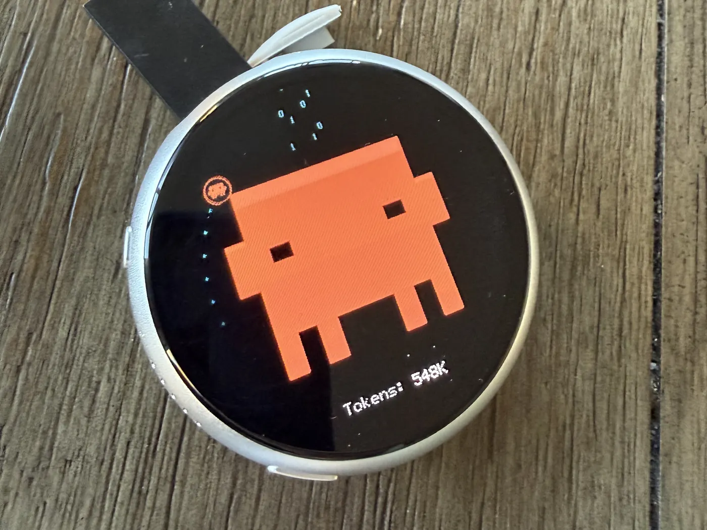 A round AMOLED board in a silver aluminium case on a wooden desk, showing an orange pixel-art character with binary digits falling beside it, a small Claude Code badge, and the text Tokens: 548K.